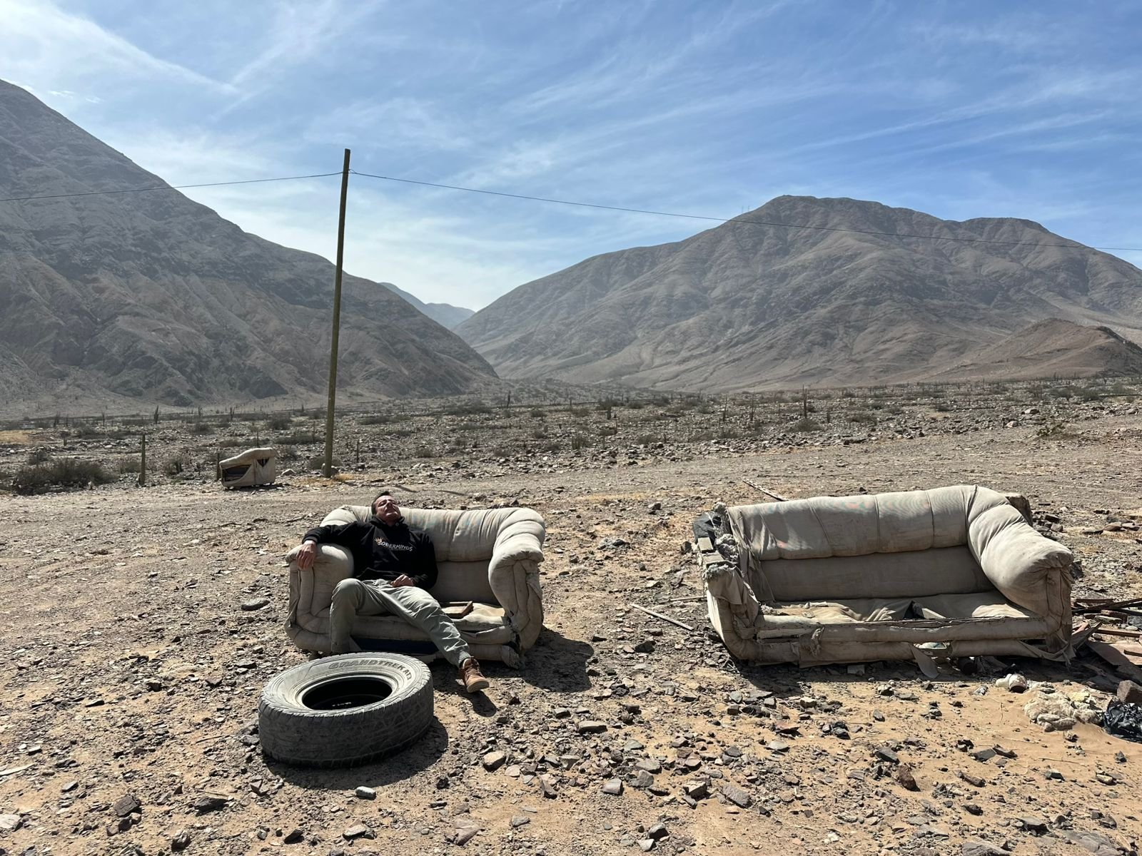 Man sitting alone on a discarded couch in an empty desert valley, facing distant mountains.