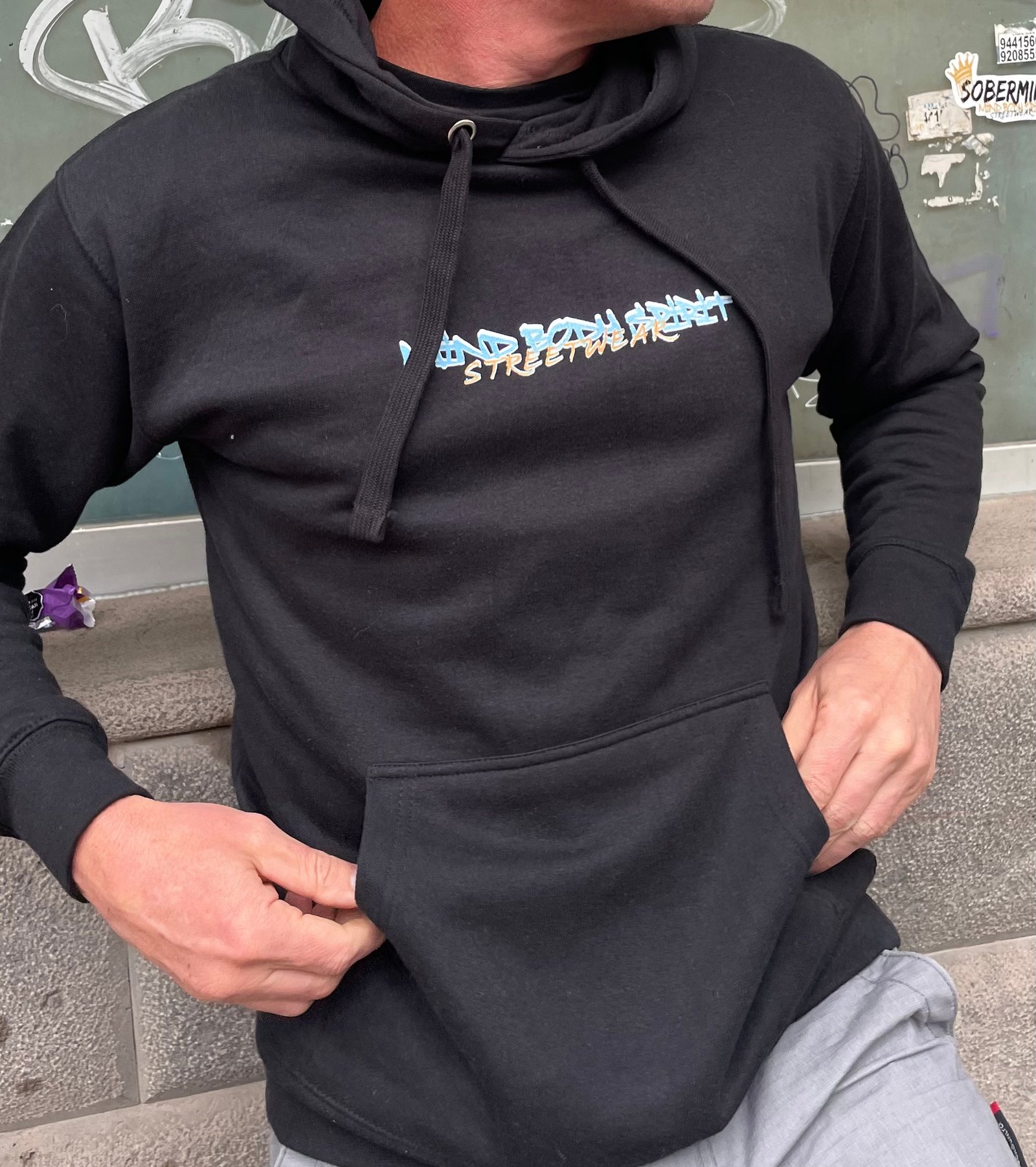 Man wearing a black Soberminds Mind Body Spirit Streetwear hoodie, front view, graffiti wall background