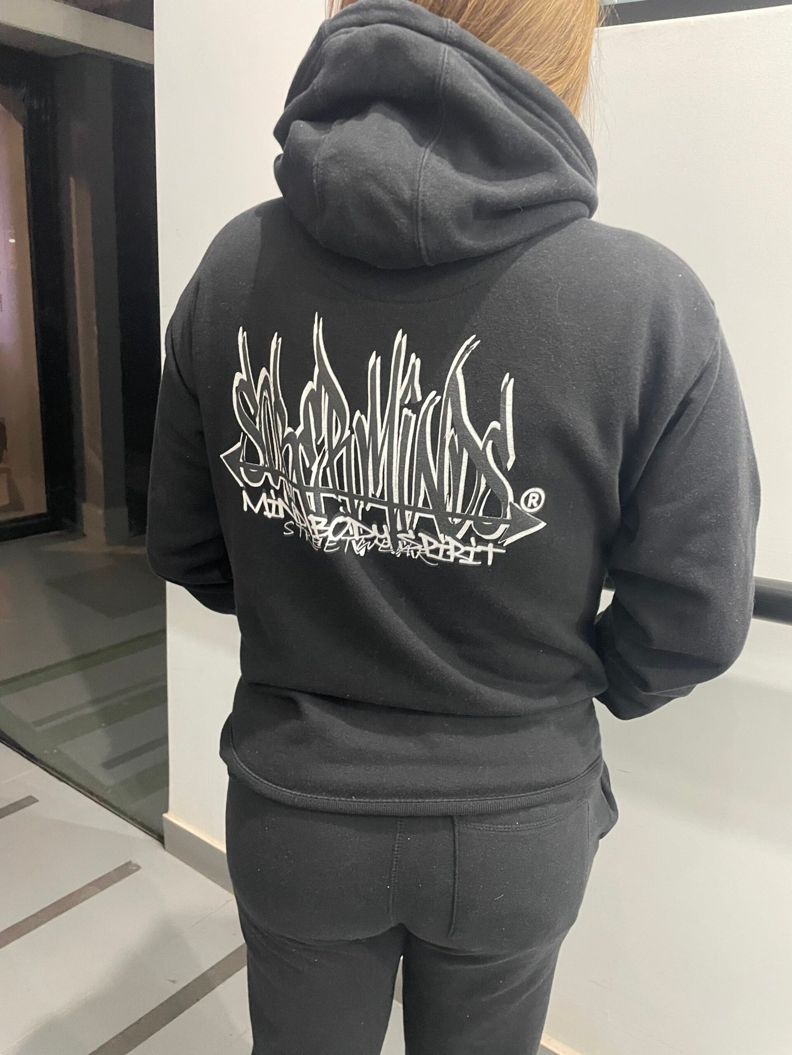 Woman wearing a black Soberminds Mind Body Spirit hoodie, back view, walking down a hallway
