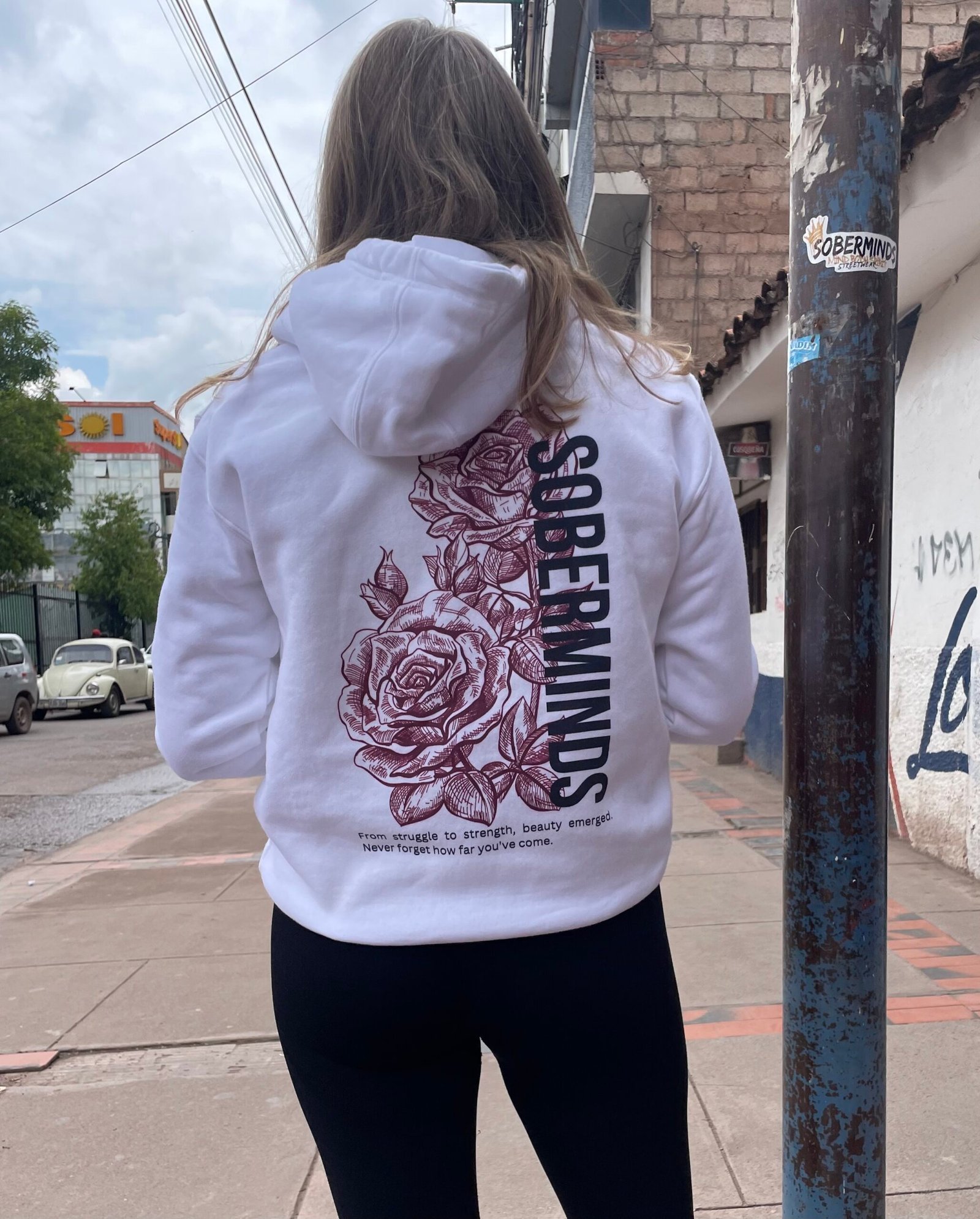 Woman walking away wearing a white Soberminds rose-design hoodie on a city street